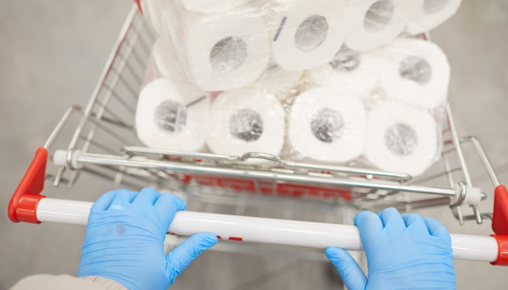 Person wearing blue gloves pushes a shopping cart filled with packs of wrapped toilet paper. The cart has red handles, conveying a sense of bulk purchase.