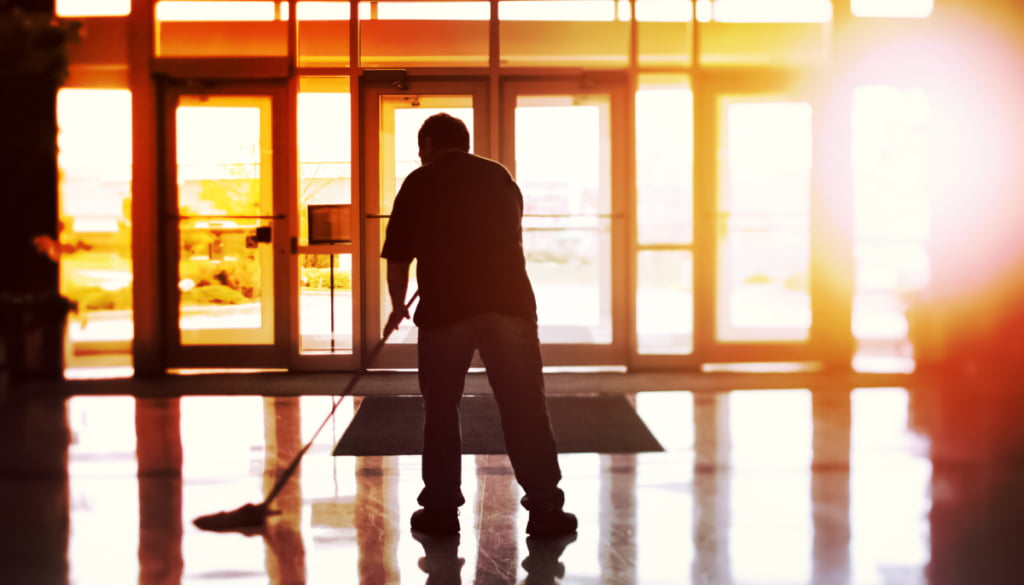 A person mops a shiny floor near glass doors at sunrise or sunset, creating a warm glow. The silhouette suggests calmness and diligence.