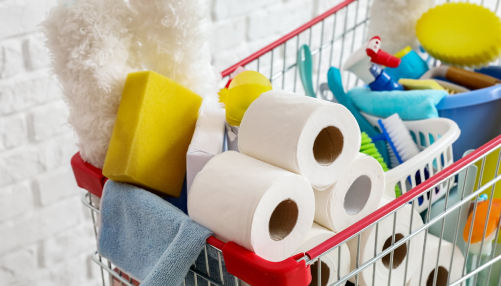 A shopping cart filled with cleaning supplies, including toilet paper rolls, sponges, brushes, and a mop, conveying a sense of readiness for cleaning.