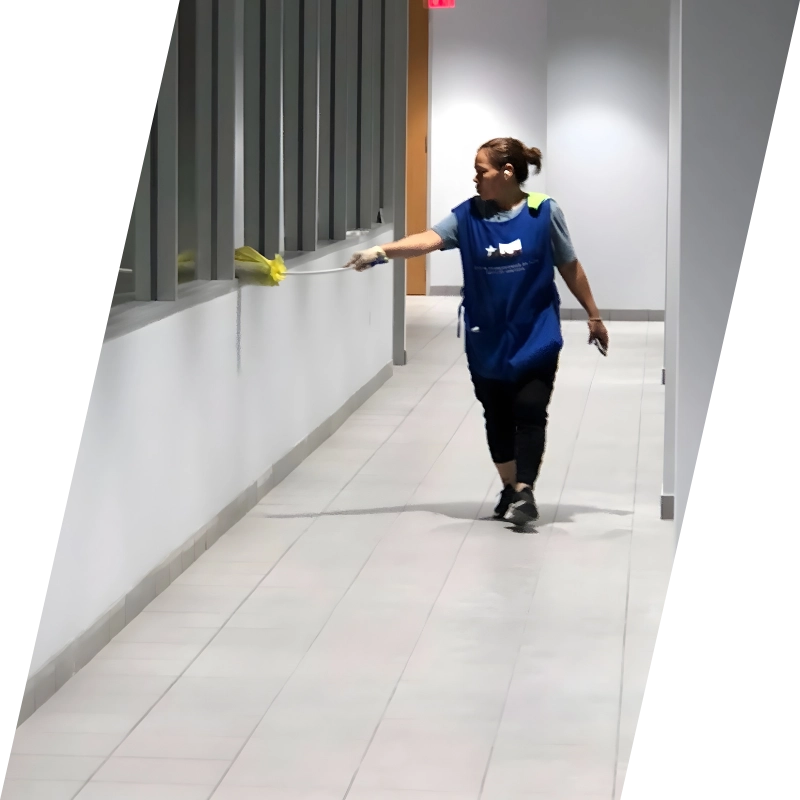 A woman in a blue apron dusts a hallway window with a yellow duster. The setting is bright and clean, conveying a sense of tidiness and focus.