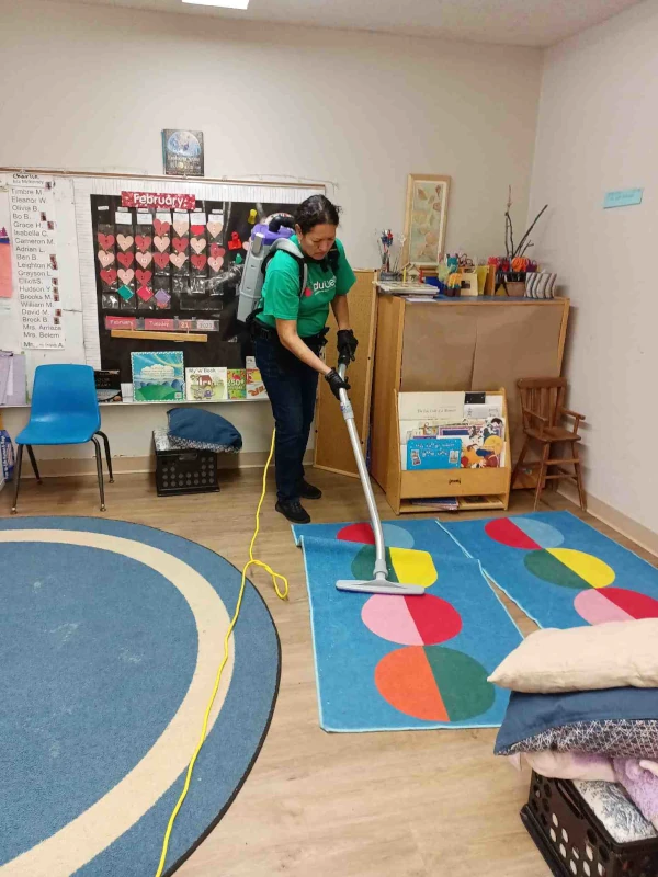 A person is vacuuming a colorful classroom rug with a backpack vacuum. The room is decorated with a heart-themed February calendar and educational materials.
