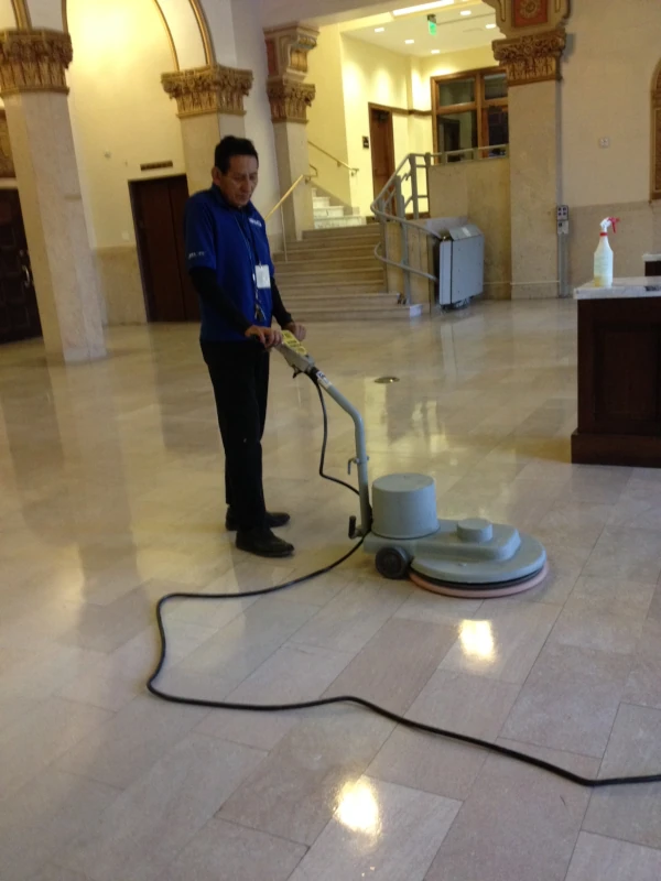 A person operates a floor polishing machine in an elegant, well-lit hall with marble columns and shiny tiles, conveying a sense of cleanliness and order.