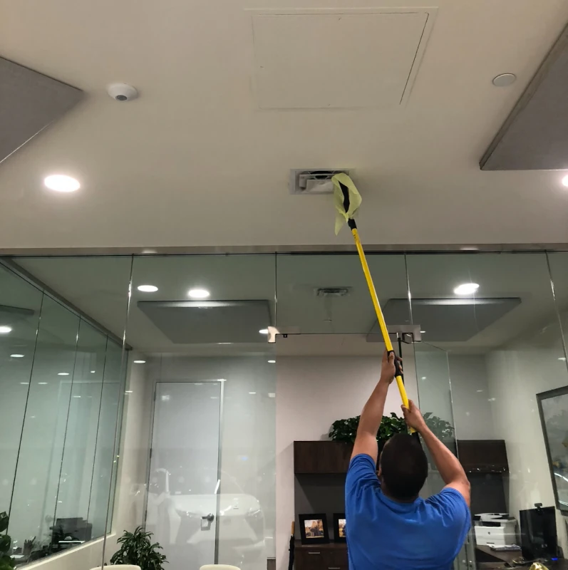 A person in a blue shirt uses a yellow pole to clean an air vent on a ceiling in a modern office with glass walls, creating a focused, tidy atmosphere.