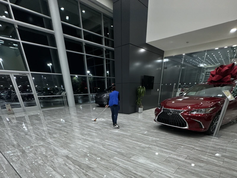 A person in a blue shirt cleans a shiny car showroom floor at night. A red car with a large bow is prominently displayed, creating a festive atmosphere.