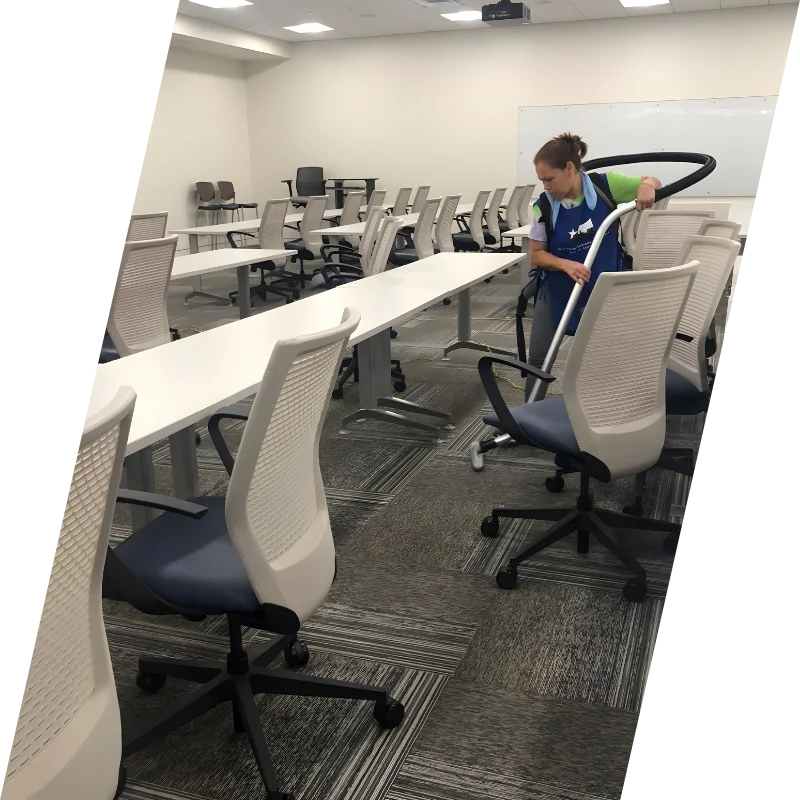 A person vacuuming an empty classroom with white tables and mesh-backed chairs. The space is organized, with a clean and focused atmosphere.