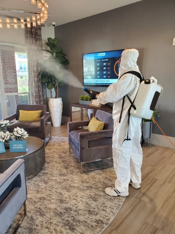 Person in protective suit sprays disinfectant in modern living room with TV displaying weather, gray chairs, yellow cushions, and plants, conveying cleanliness.
