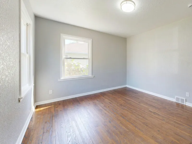 Empty room with wooden floor, gray walls, and a single window letting in natural light. The ceiling light is on, creating a bright and airy atmosphere.