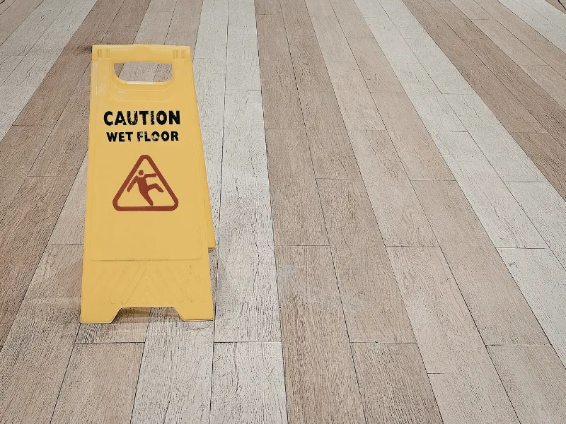Yellow "Caution Wet Floor" sign on light wood flooring, emphasizing safety with a slipping figure icon. The setting is clean and orderly.