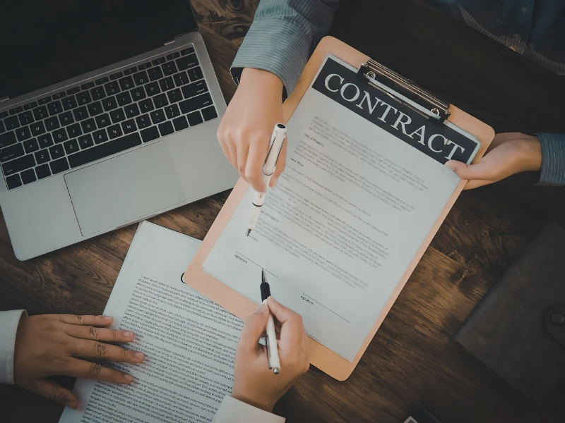 Overhead view of a contract being signed on a clipboard, with two hands holding pens. A laptop is nearby. The scene conveys a professional setting.