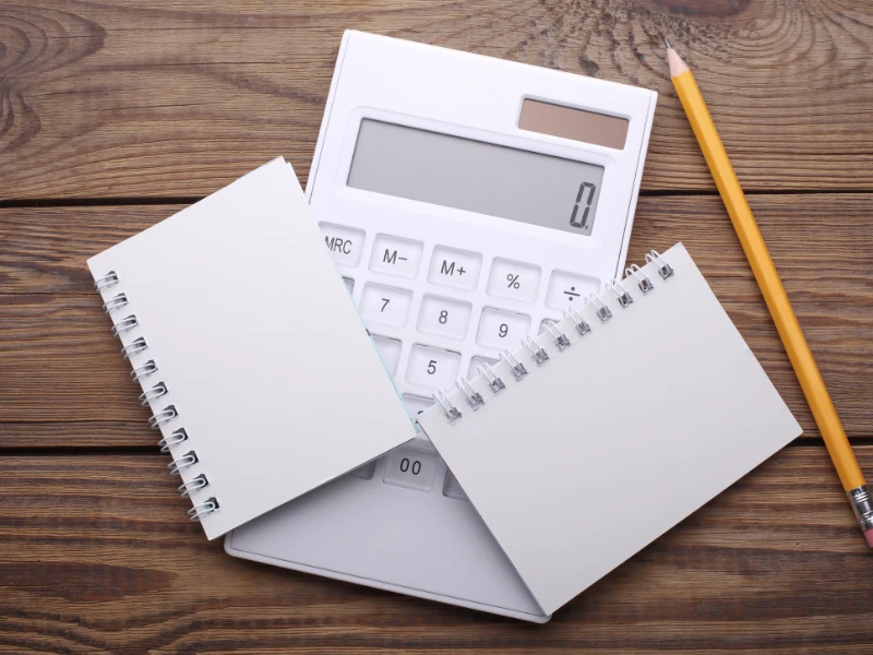 A large white calculator displays zero, flanked by two small spiral notepads and a sharpened pencil on a wooden desk, conveying a sense of readiness.