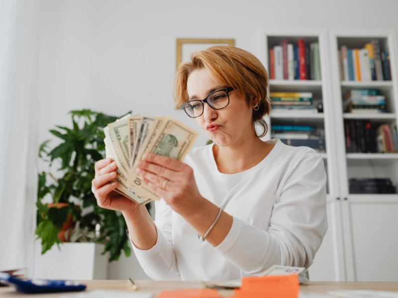 A woman in glasses, with a focused expression, counts a stack of US dollar bills. She sits at a desk, with bookshelves and plants in the background.