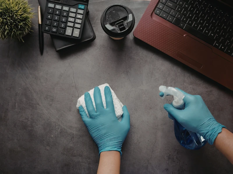 Hands wearing blue gloves clean a desk with a cloth and spray bottle. Nearby are a laptop, calculator, notebook, and coffee cup, conveying cleanliness.