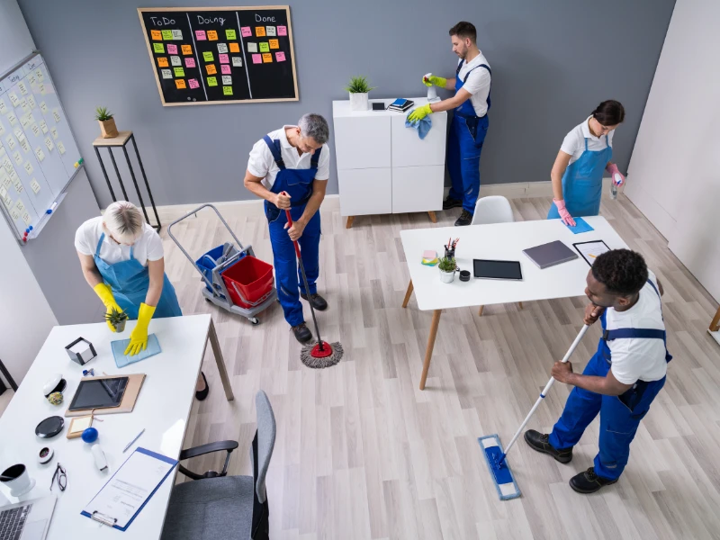 A team of five janitors in blue uniforms cleans an office. They mop the floor, dust, and wipe surfaces. A task board and desks with laptops are visible.