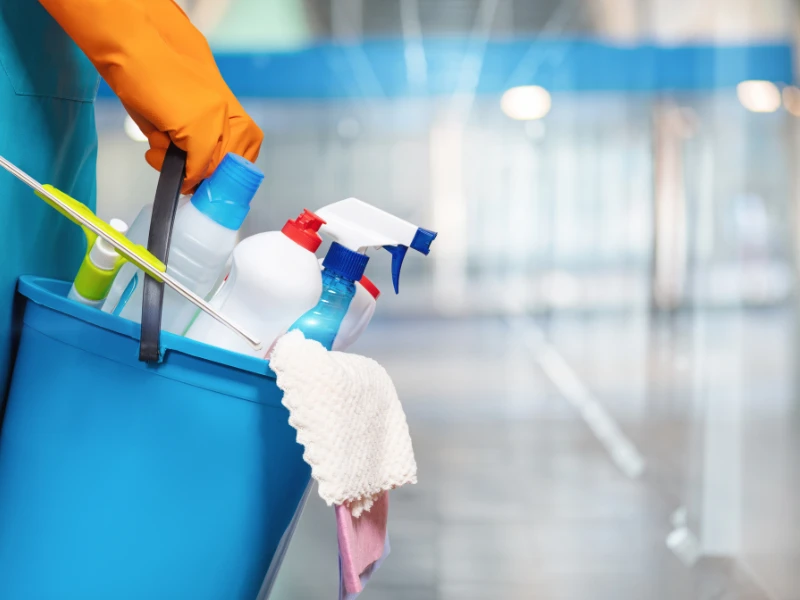 A person wearing an orange glove holds a blue bucket filled with cleaning supplies, including spray bottles and cloths, in a bright, modern hallway.