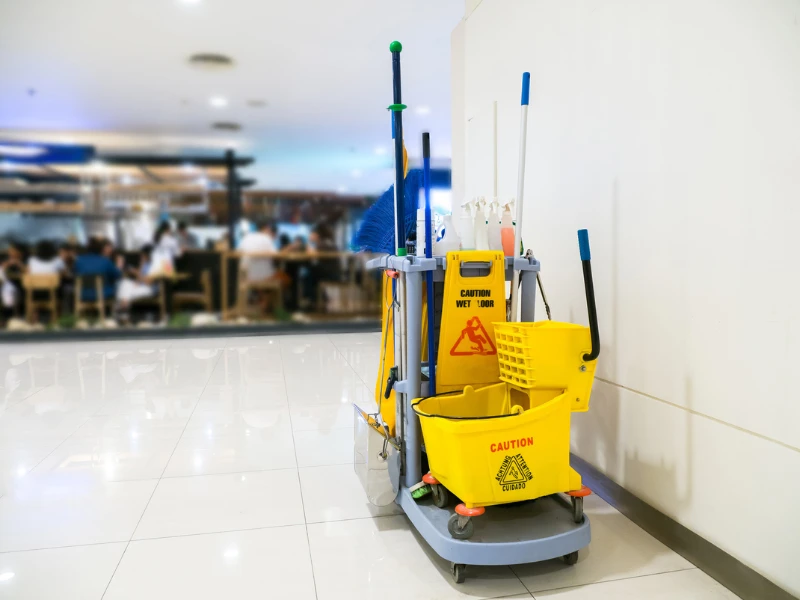Cleaning cart with mops, brooms, and a yellow "Caution Wet Floor" sign stands in a bright hallway, with blurred restaurant scene in the background.