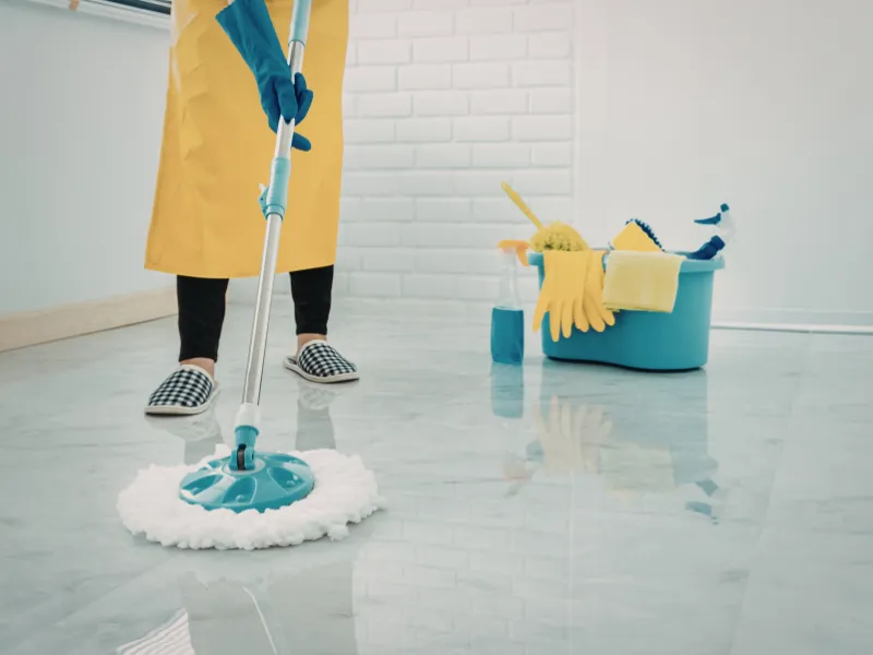 Person in a yellow apron and gloves mops a shiny tiled floor. A bucket with cleaning supplies is nearby, conveying a clean and tidy atmosphere.