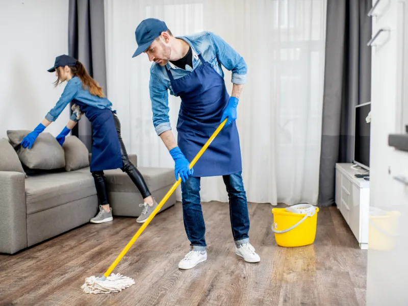 Two people in blue aprons and gloves clean a modern living room. One mops the wooden floor near a yellow bucket, while the other wipes a gray sofa.