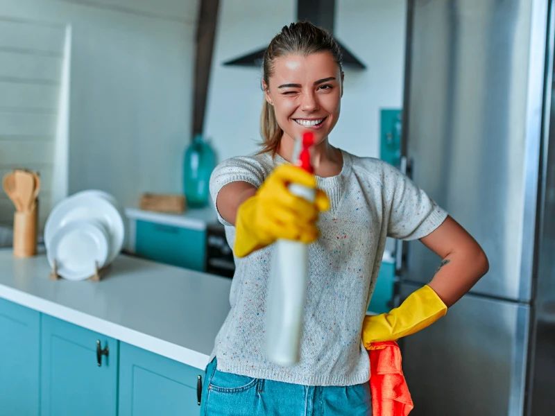 A smiling woman in yellow gloves holds a spray bottle forward, winking playfully in a clean, bright kitchen. She appears cheerful and ready to clean.